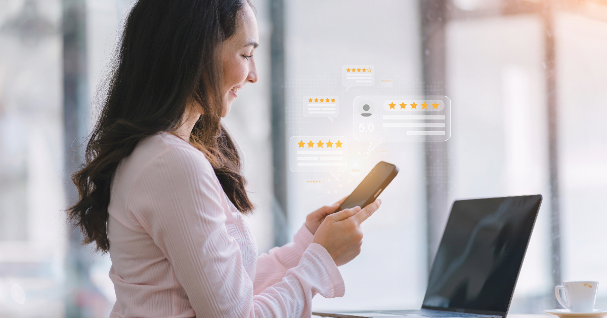 A woman in a light pink shirt smiles while using a smartphone, with a laptop on the table in front of her. User rating icons and review bubbles are digitally overlaid in the background, capturing an elevated customer experience through her personalized shopping assistant on an e-commerce platform.