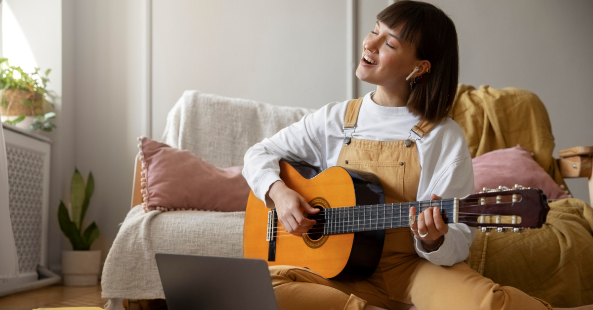 A person wearing overalls plays an acoustic guitar while sitting on the floor in a cozy, well-lit room with heavy curtains, enjoying acoustic comfort, and a laptop in front of them.