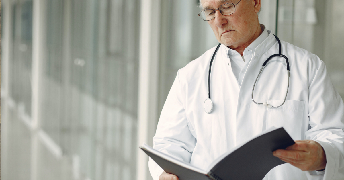A doctor in a white coat and stethoscope reads a treatment report in a bright, modern hallway.