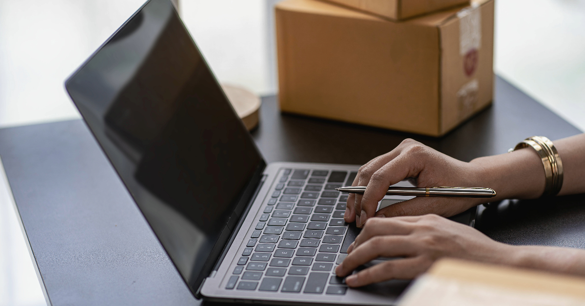 Person's hands typing on a laptop keyboard, with cardboard boxes in the background, likely working on e-commerce operations and inventory optimization.