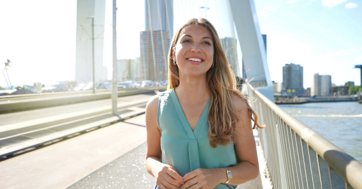 A woman with long hair and a teal sleeveless top stands on a bridge in a city, smiling with buildings and water in the background, perhaps considering ways to make extra income, like selling feet pics.