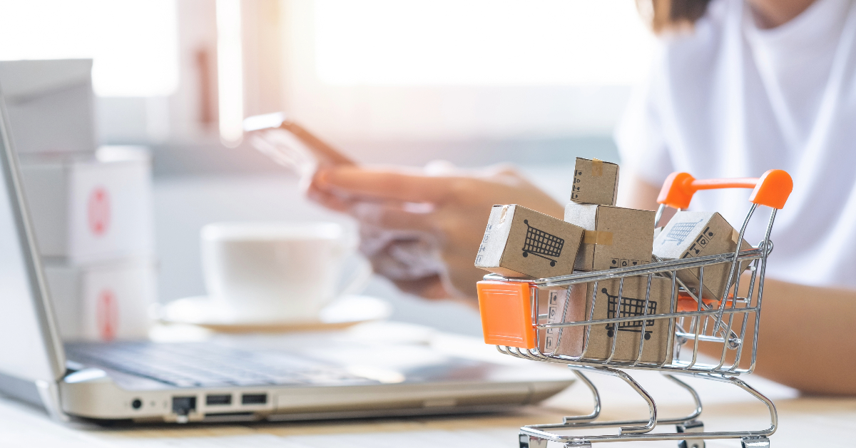 A person using a smartphone next to a laptop, with a mini shopping cart containing small cardboard boxes in the foreground—an ideal setup for SEO optimization and lead generation activities.