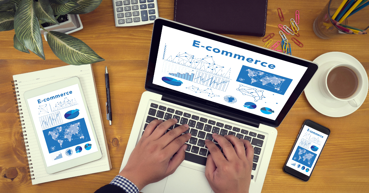 A person types on a laptop displaying e-commerce data charts. A tablet, smartphone, notepad, plant, coffee cup, calculator, and colorful paperclips are also on the wooden desk. SaaS tools help manage these insights efficiently.
