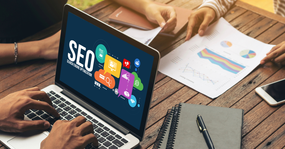 A person types on a laptop displaying an SEO screen. Two others review papers and charts at the wooden table, discussing e-commerce strategies. Notebooks and a pen are also on the table.