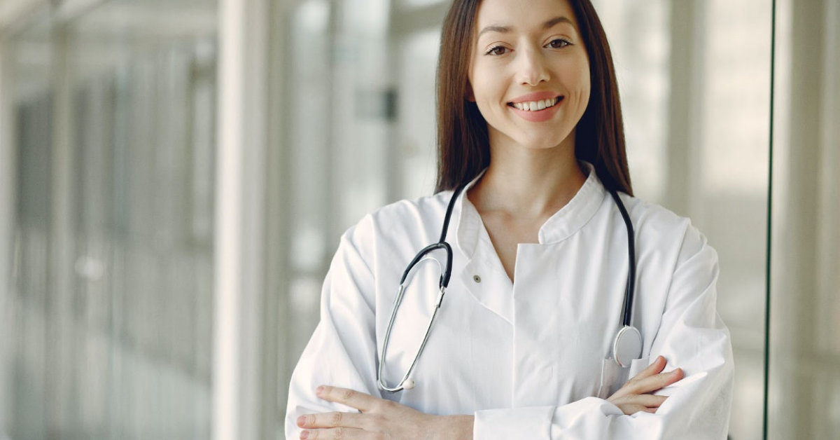 A woman in a white coat with a stethoscope around her neck stands with arms crossed and smiles. She is in a bright, modern indoor setting, exemplifying the expertise involved in Hospitalist Billing.