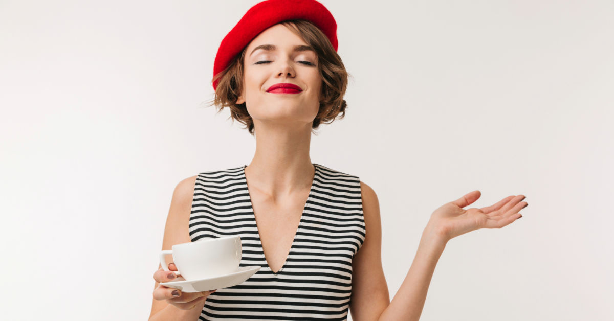 A woman wearing a red beret, black-and-white striped dress, holds a white teacup and saucer in one hand and gestures with the other. With a content expression on her face, she looks as if she's enjoying a serene day in France.