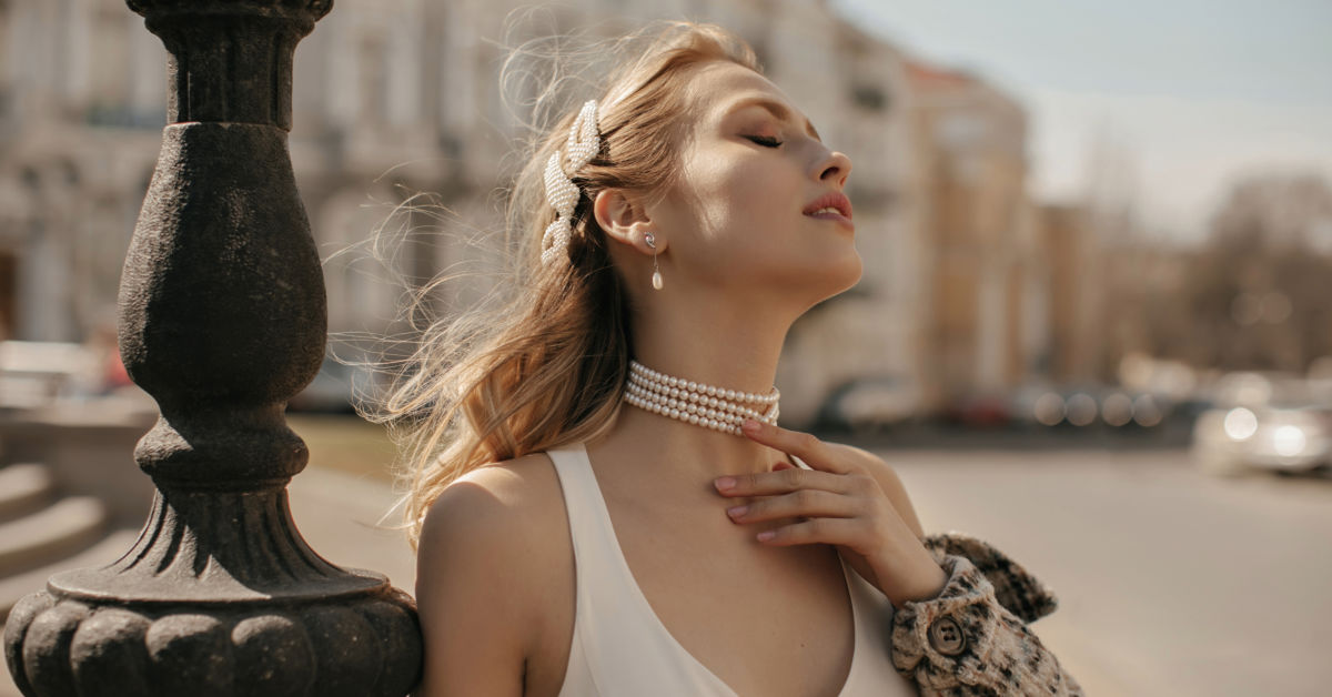 Woman with closed eyes, wearing pearl jewelry and a white top, stands beside a lamp post on a sunny day in an urban setting in Germany.