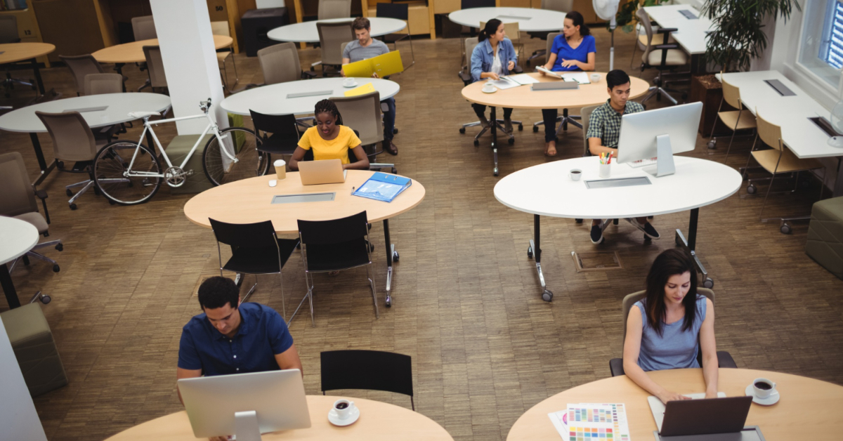 A group of people working on computers and laptops in an open, modern coworking space with several round tables and a bicycle against the wall, showcasing why it's the preferred choice for professionals in 2024.