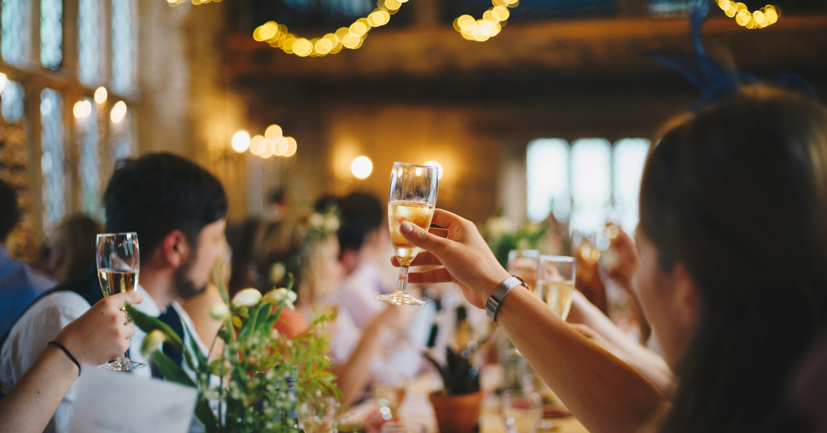 People raising glasses in a dimly lit room decorated with string lights, seated around a long table adorned with flowers and greenery, creates the perfect setting that any wedding planner would dream of to win over customers.