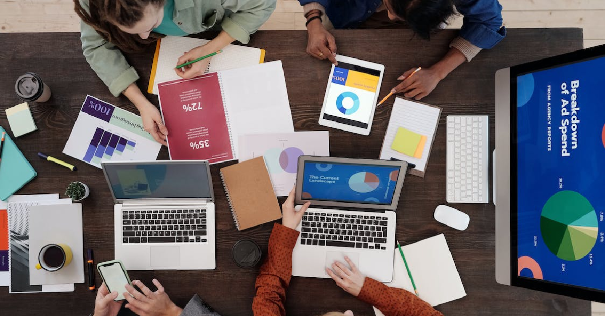 Overhead view of a workspace with five people working on laptops, tablets, and documents featuring graphs and charts. Several notebooks, sticky notes, and coffee cups are also on the table as they discuss Clean and Reliable Data.