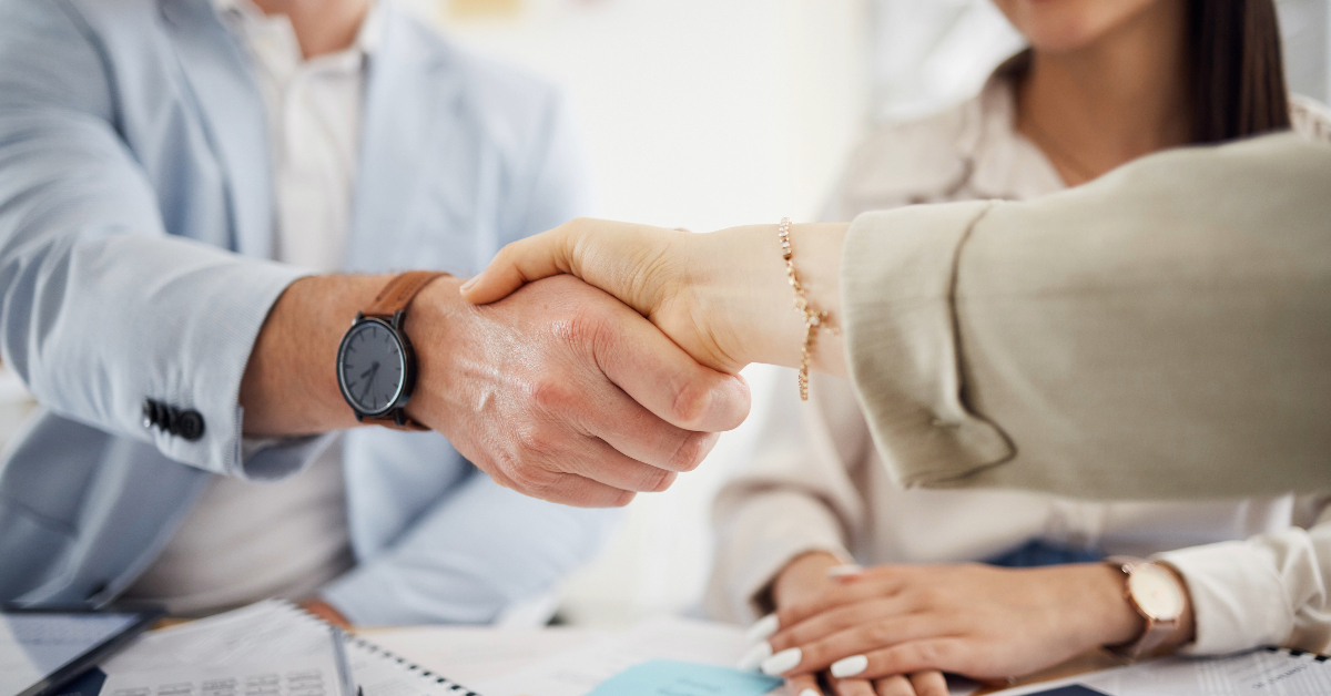 Two people shaking hands over a desk filled with office supplies and documents, with another person from Hero Digital sitting nearby, possibly discussing the B2B Buyer Report.
