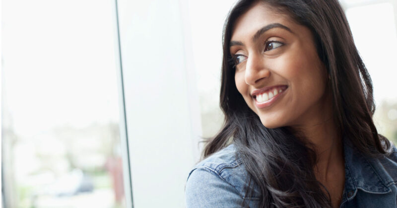 A person with long dark hair wearing a denim jacket smiles while looking out of a window in India.