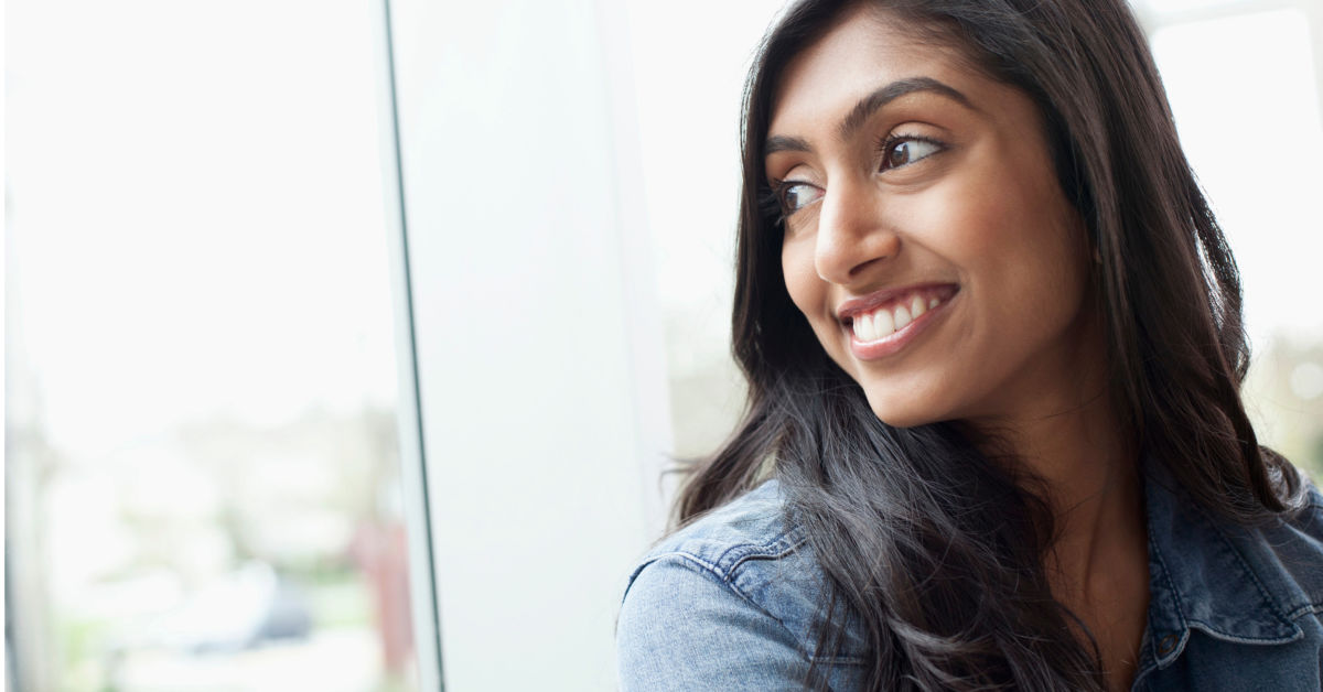 A person with long dark hair wearing a denim jacket smiles while looking out of a window in India.