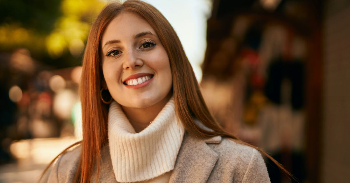 A woman with long red hair, wearing a beige coat and white turtleneck, smiles outdoors on a sunny day in Ireland.
