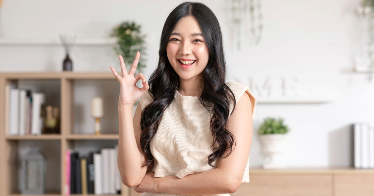 A woman with long dark hair stands indoors, smiling and making an "OK" hand gesture with her right hand. She is wearing a sleeveless beige top and has a bookshelf in the background, adorned with various books about Japan.