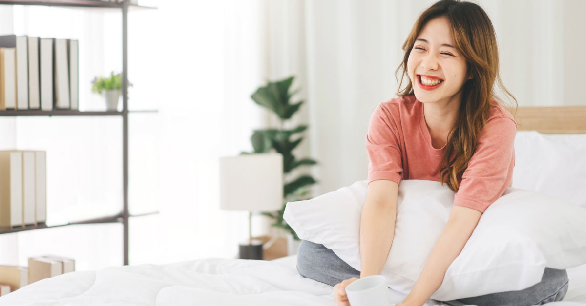 A woman sits on a bed in South Korea, smiling and holding a cup. She wears a pink shirt and blue jeans. Bookshelves and a potted plant are in the background.