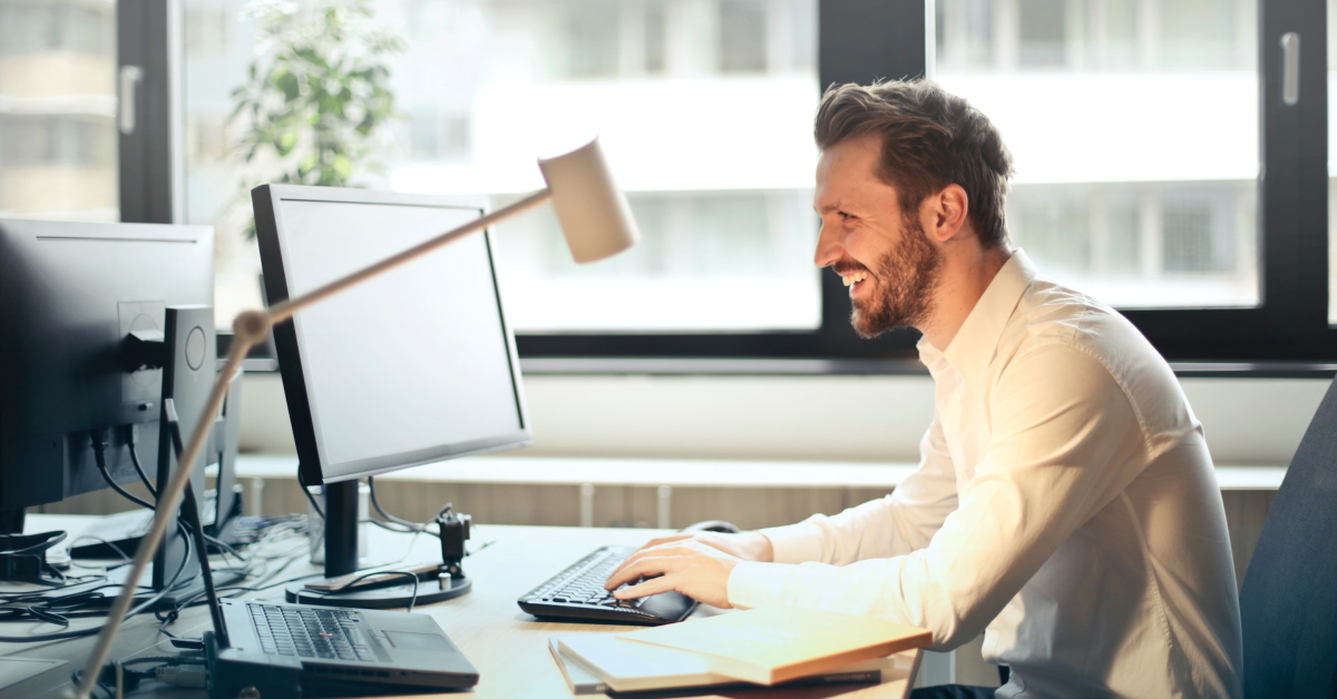 A man in a white shirt is sitting at a desk, smiling and typing on a keyboard in front of a computer monitor. The desk has books, an email management guide, a lamp, and another monitor in the background.