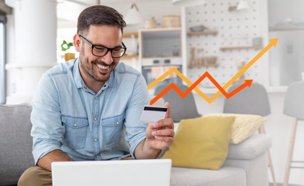 A man sits on a couch holding a credit card and smiling at his laptop, with an upward-trending orange graph displayed in the background, reflecting the success of E-commerce optimisation.