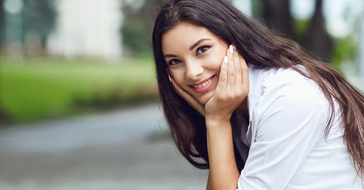 A woman with long dark hair smiles while resting her chin on her hands, outdoors with a blurred green background. She is wearing a white blouse and enjoying the sunny weather in Mexico.