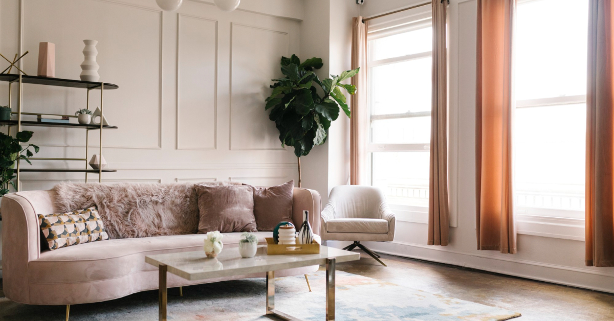 A living room features a pink sofa, patterned cushions, a coffee table, an armchair, a large indoor plant, a wall-mounted shelf with decor items, and large windows with light brown curtains. Transform your space into your dream room with these elegant touches.