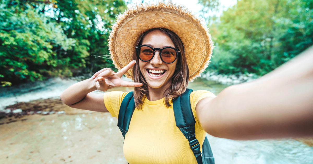 A person wearing a straw hat and sunglasses smiles while making a peace sign with one hand. They are outdoors next to a wooded area in New Zealand.