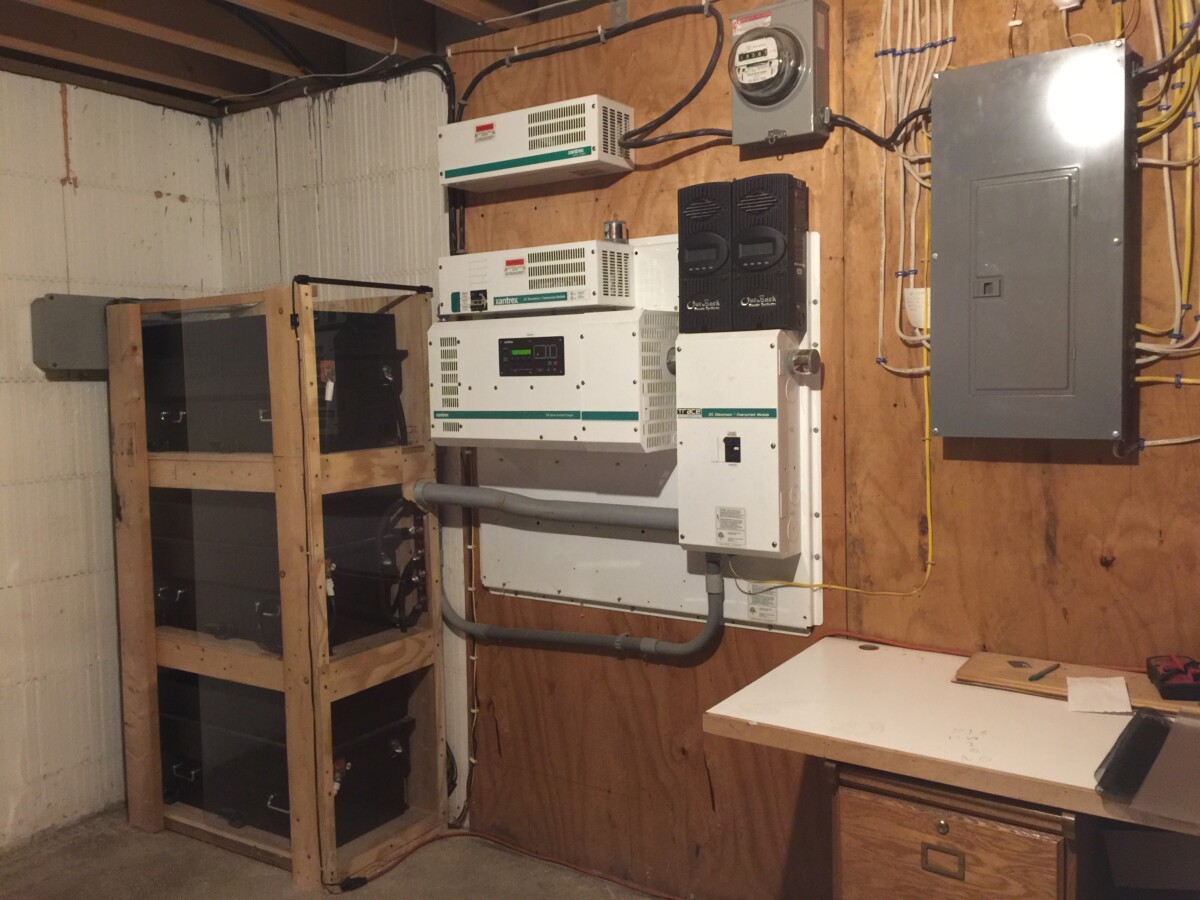 Basement utility room featuring electrical equipment mounted on the wall, including circuit breakers, inverters, and a comprehensive Battery Energy Storage system stored on a wooden rack. A table is seen on the right side.