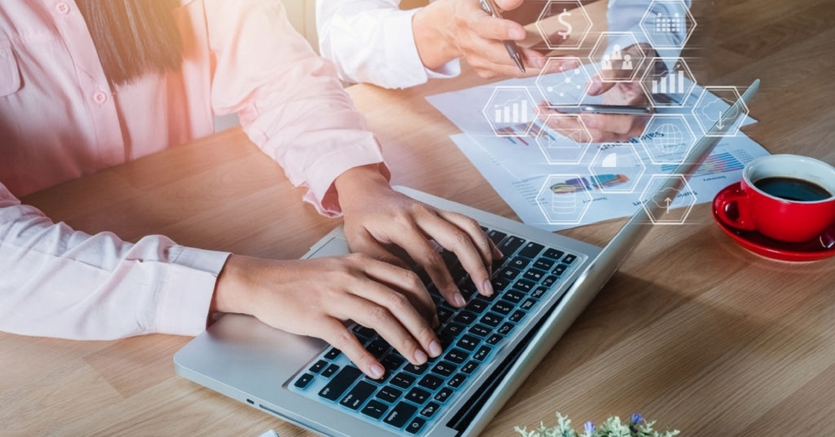 Two people are working at a table with a laptop and documents, discussing effective tricks for boosting sales. One is typing, while the other points at charts on paper. A red coffee cup is placed beside them, and holographic icons add a futuristic touch as they plan their eCommerce site strategy.