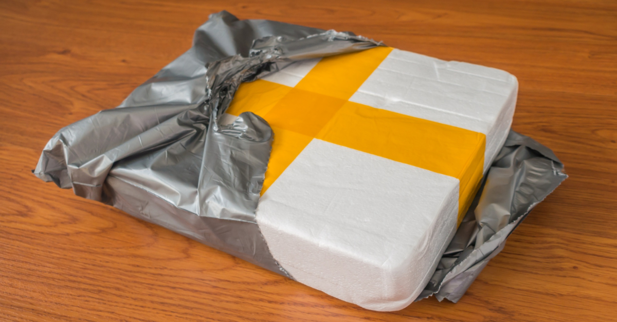 Partially opened grey plastic packaging reveals a white foam container with yellow tape on a wooden surface.