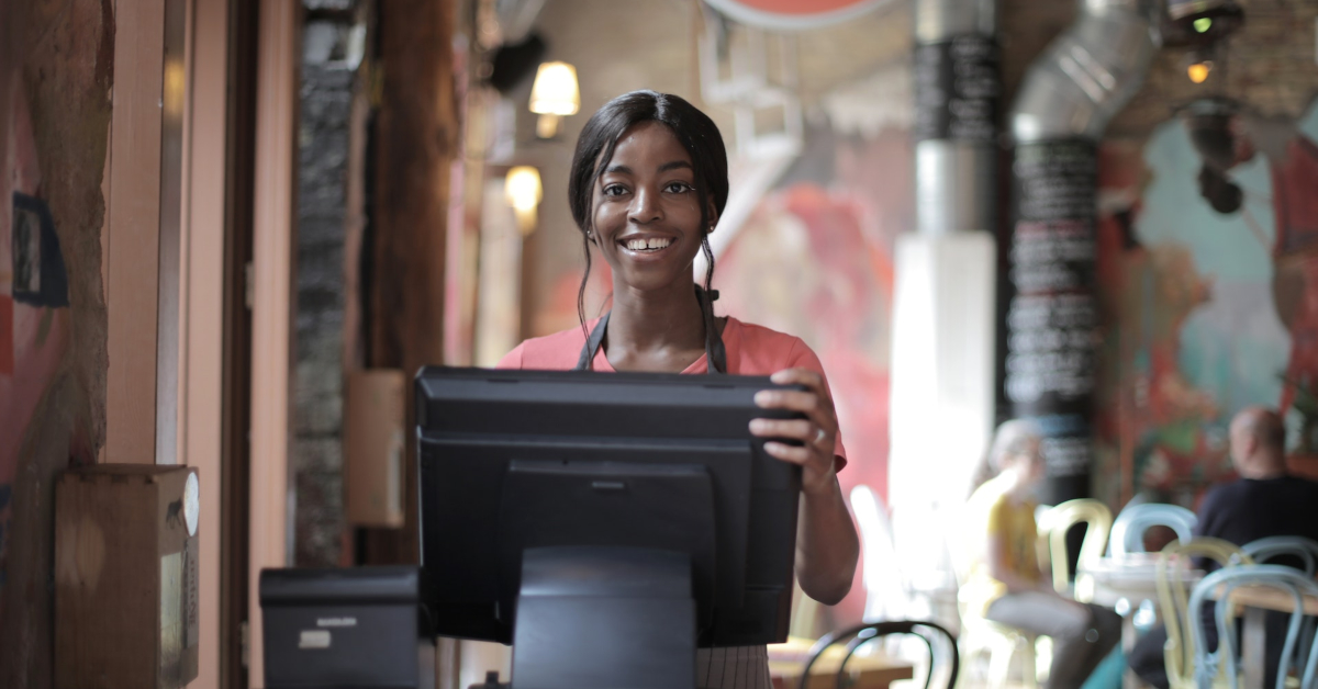 A person stands behind a cash register at a small business cafe, smiling. The background shows tables and colorful decor, creating an inviting atmosphere—perfect for learning how to please your customers.