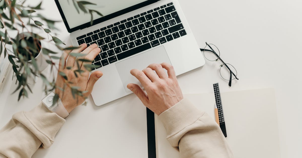 A person wearing a beige sweater types on a silver laptop. Nearby are a pair of eyeglasses, a plant, and an open notebook with a pen on a white desk, creating a serene space ideal for focusing on recovery and rebuilding your life after any workplace injury.