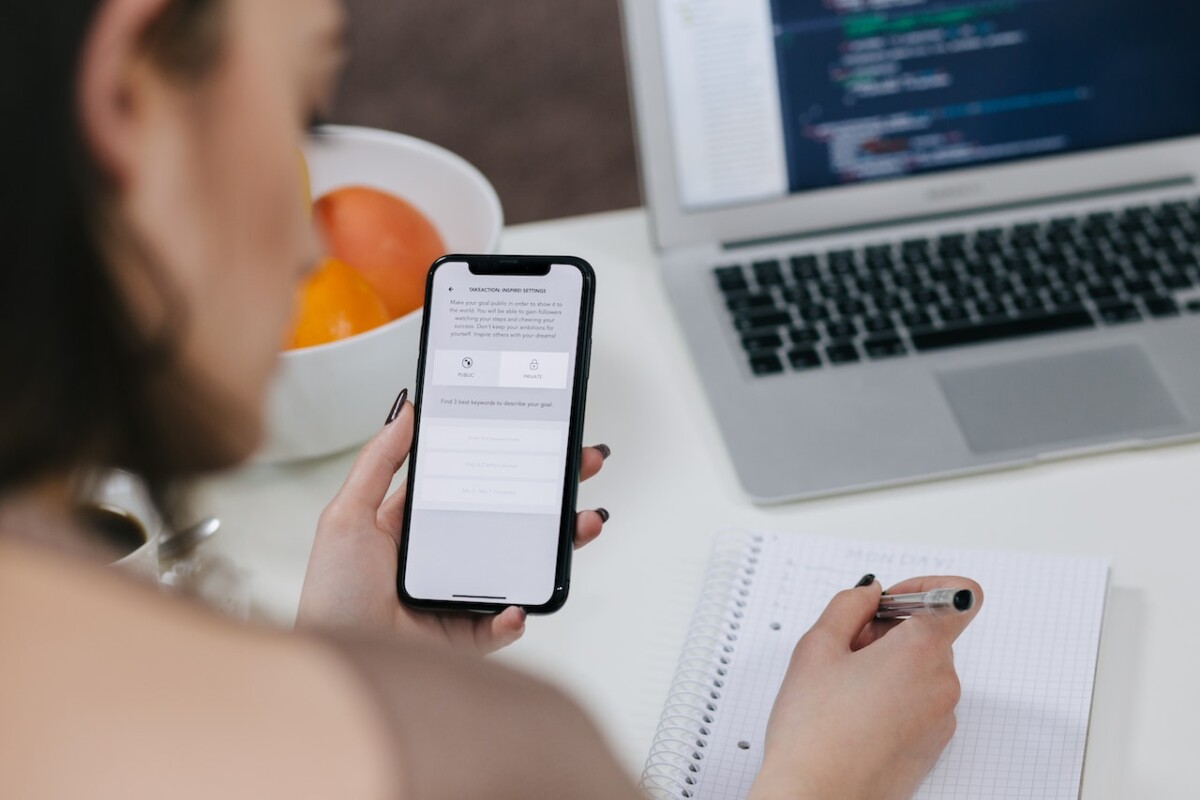 Person using a smartphone and taking notes on a notepad with a laptop displaying code on the desk nearby, researching SEO services in the UK.