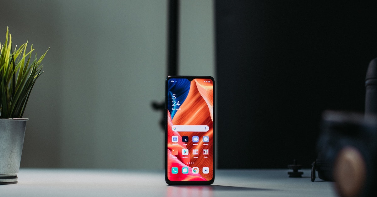 A mobile phone with a colorful, abstract design on its screen stands on a table next to a potted plant and a camera, making it an aesthetic choice for any tech enthusiast.