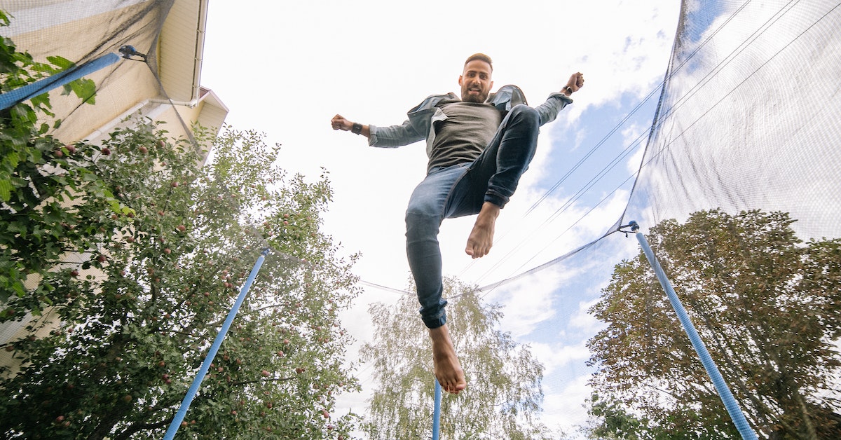 A man in casual clothing jumps on a trampoline surrounded by trees and a house under a partly cloudy sky, enjoying bouncy fun.