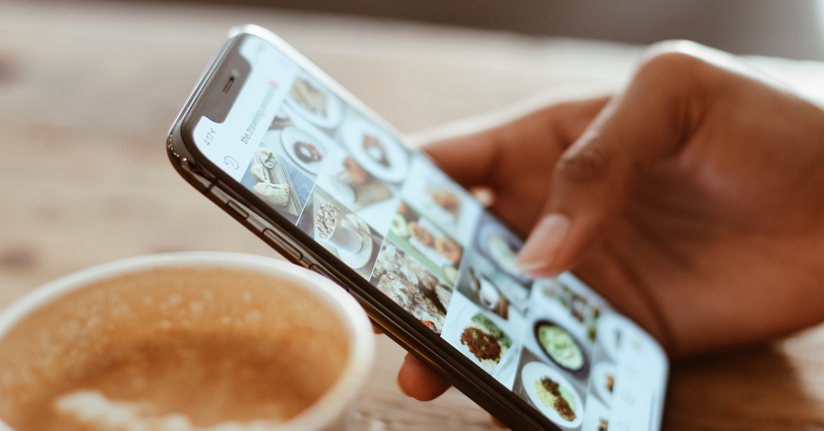 A person browsing through food photos on a smartphone while holding a latte in a cup on a wooden table, creating authentic connections in the digital world.