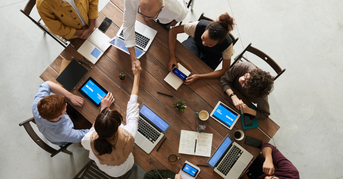 A top-view photo of seven people from various business departments sitting around a table with laptops and tablets, shaking hands, with notebooks, a coffee cup, and potted plants on the table.