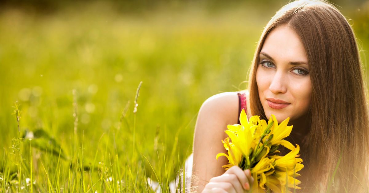 A person with long brown hair lies in a field of green grass, holding a bouquet of yellow flowers, in the countryside of Russia.