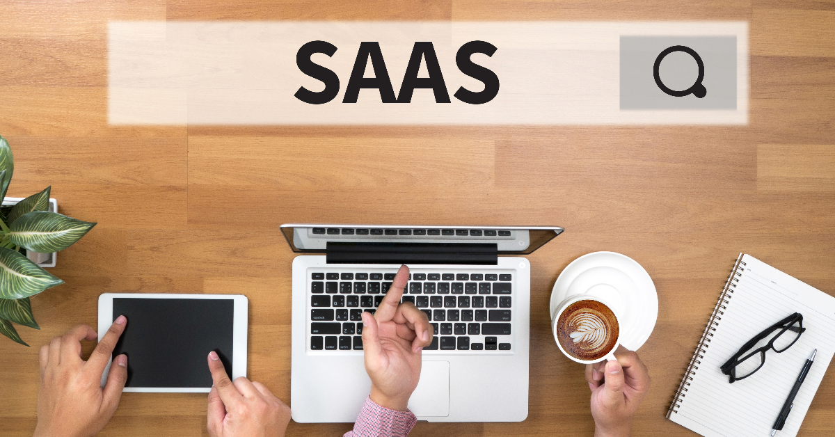 Overhead view of a workspace with a laptop, tablet, coffee cup, notebook, pen, and plant. The word "SAAS" and a search icon are displayed above the workspace. Three hands point to different items, illustrating competitor strategies in an SEO-optimized environment.