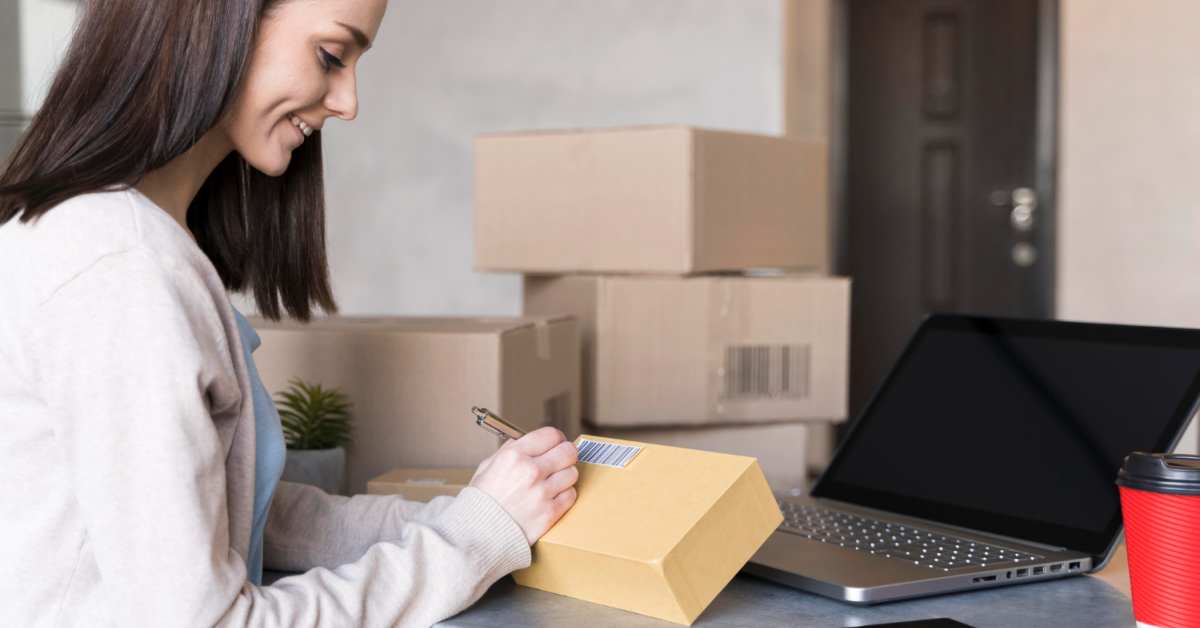 A woman sits at her desk, writing on a shipping label for a small box. Surrounded by cardboard boxes, a laptop, and a red coffee cup, she's immersed in her work—creating custom mailer boxes that elevate her brand with personalized packaging.
