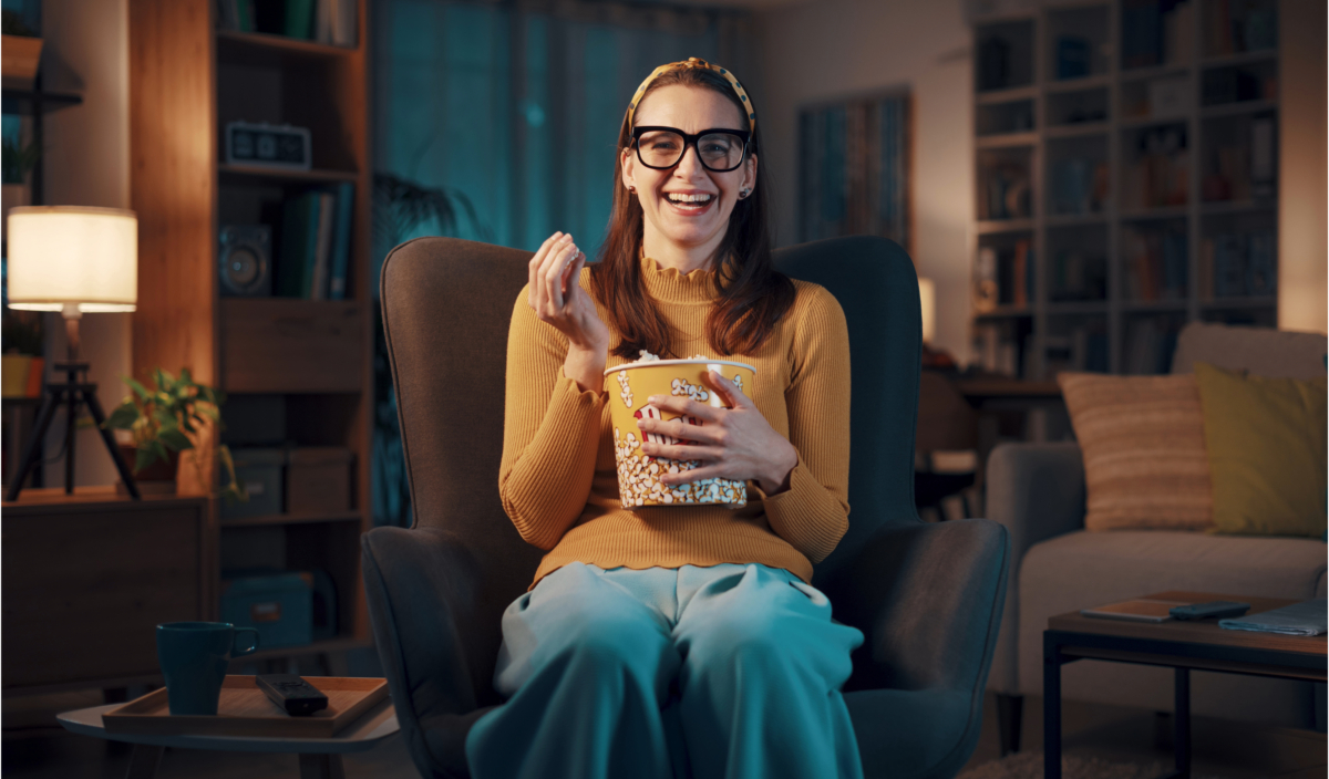 A person wearing glasses and a headband is sitting in a comfortable chair, smiling, and eating popcorn from a large bucket while enjoying unlimited streaming in a cozy, dimly-lit living room.