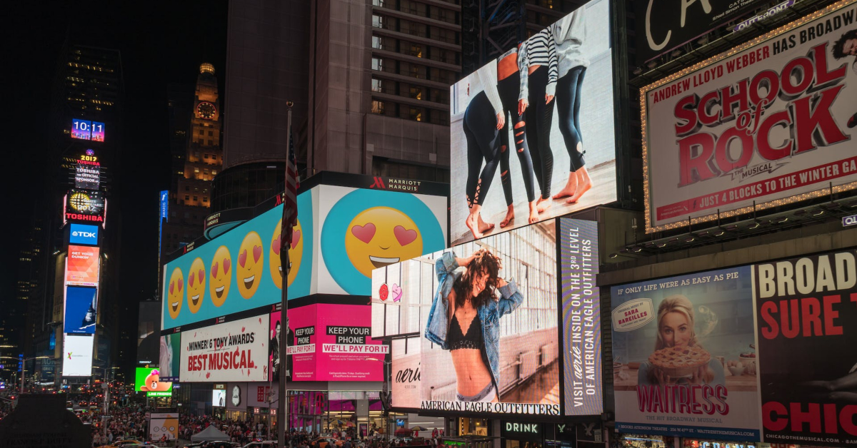 Times Square at night featuring brightly lit outdoor billboards advertising musicals, fashion, and emojis. People walk below, surrounded by the vibrant glow of the towering advertisements. The scene feels like a place where you could power up your experience with the latest trends.