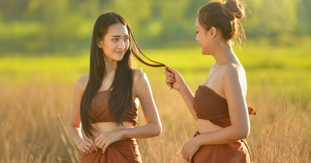 Two women in a grassy field, wearing brown traditional attire from Thailand. One woman is playfully pulling the other's hair while they both smile.