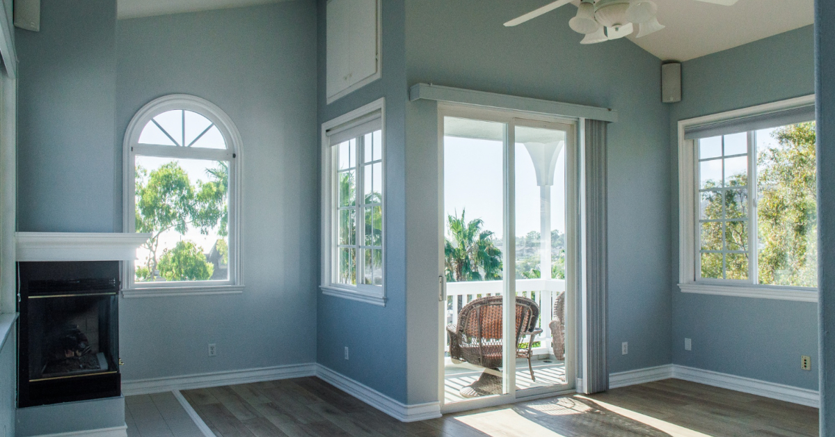 A bright room with light blue walls, a fireplace, arched windows, and sliding glass doors opening to a balcony with outdoor furniture and trees in the background showcases the benefits of replacement windows and doors.