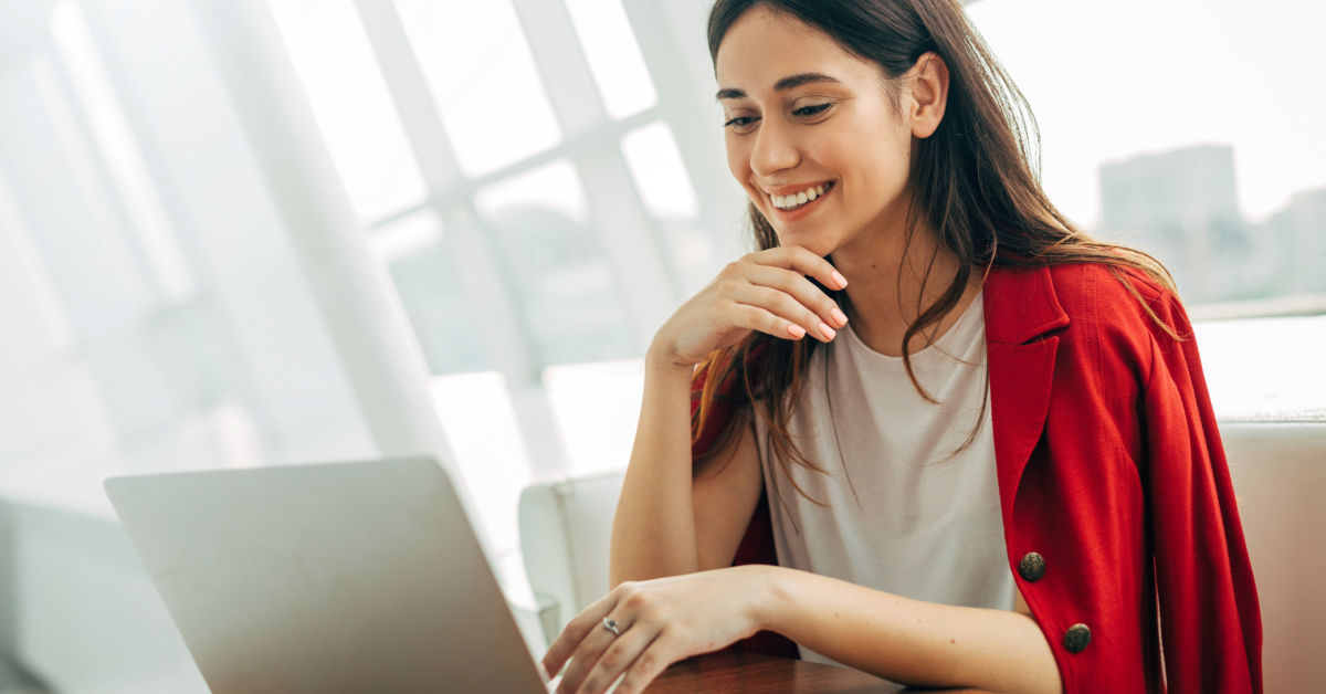 A woman wearing a red blazer is sitting at a table in Turkey, smiling while looking at her laptop.