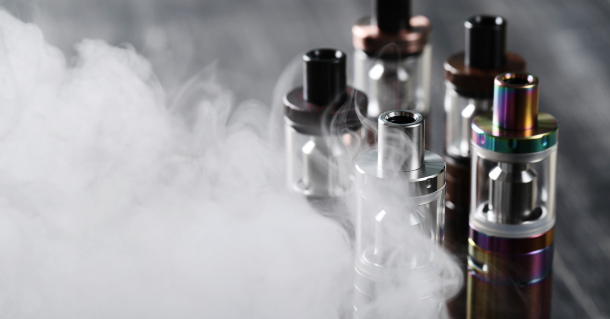 Several vape atomizers from the best disposable vape manufacturers release smoke on a dark surface, arranged closely together.