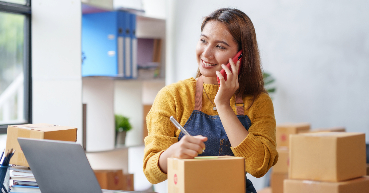 A woman is talking on the phone while writing on a notepad. She is sitting at a desk with a laptop and several cardboard boxes, likely discussing inventory management for her e-commerce business.