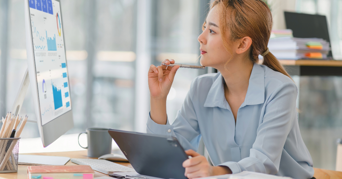 A person in a light blue shirt is sitting at a desk, holding a pen while looking thoughtfully at a computer screen displaying graphs and charts from their Growth Playbook. A tablet, a coffee cup, and notebooks are on the desk, capturing the essence of strategizing for success during the holiday season.