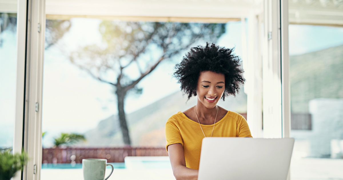A person sits at a desk, working on a laptop in a bright room with large windows. Exploring PBNs, they sip from a coffee mug while concentrating on boosting website rankings. A tree is visible outside.
