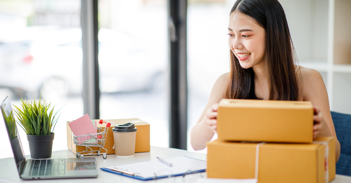 A woman smiles while handling a cardboard box at a desk with a laptop, documents, and a shopping cart filled with items, symbolizing e-commerce engagement. A coffee cup and potted plant add to the workspace's charm.