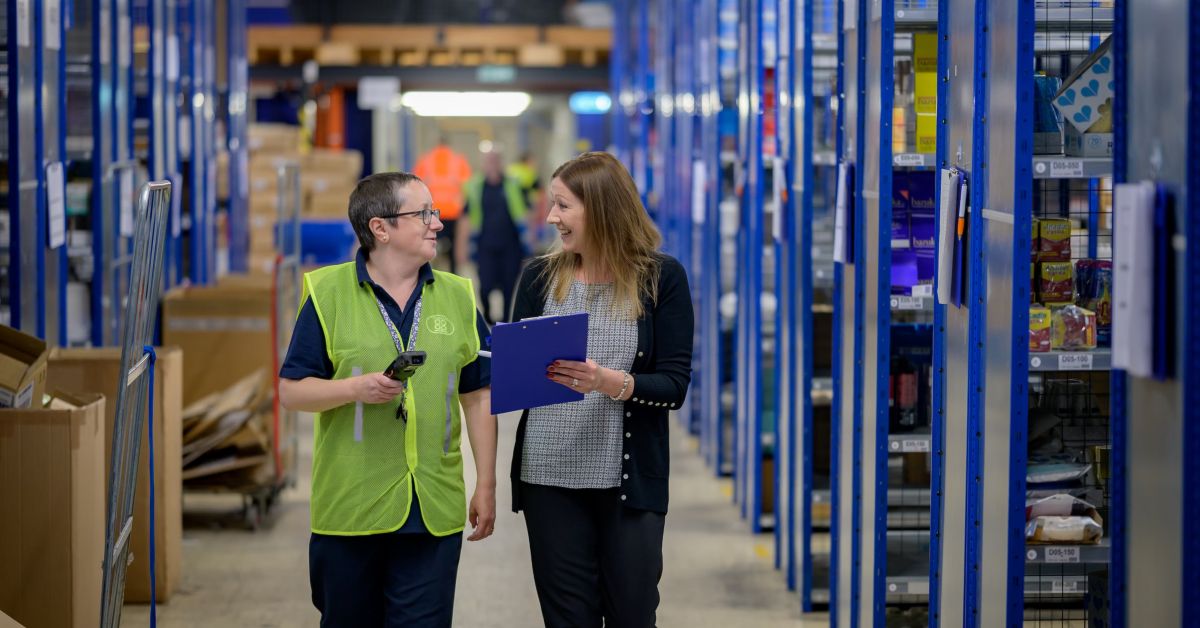 Two people walking and discussing in a warehouse, one in a high-visibility vest with a barcode scanner, the other holding a clipboard. Shelves stocked with various items line both sides of the aisle, as they examine how warehouse automation is reshaping ecommerce logistics.