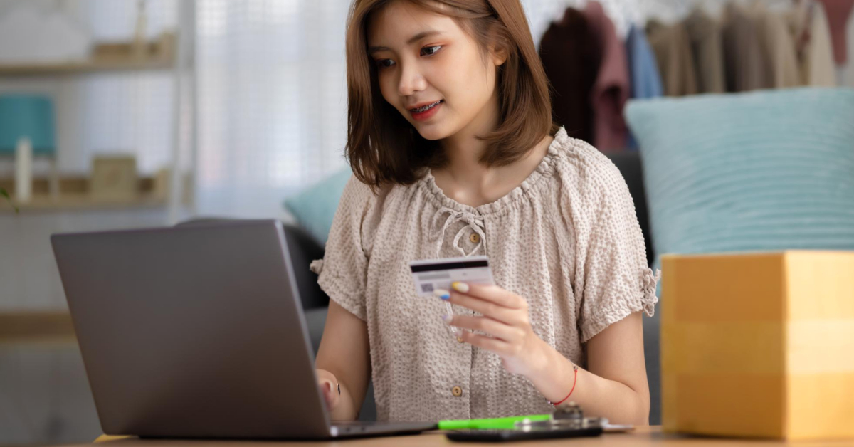 A woman sits at a desk using a laptop while holding a credit card, optimizing her ecommerce experience by browsing product pages.